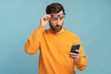 Man in orange sweater looks surprised at his phone, adjusting glasses.