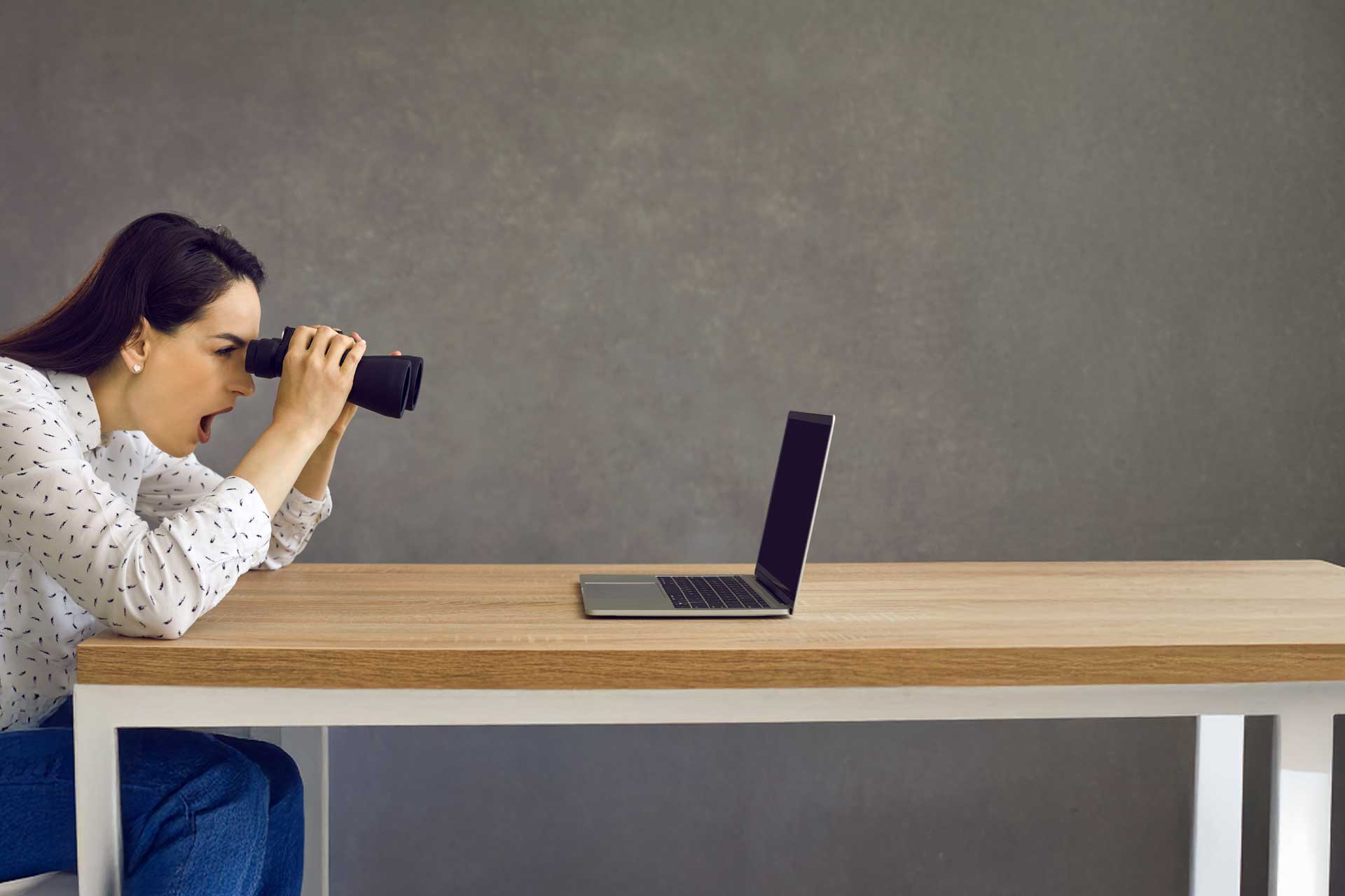 Person using binoculars for SEO analysis on a laptop screen at a wooden table.