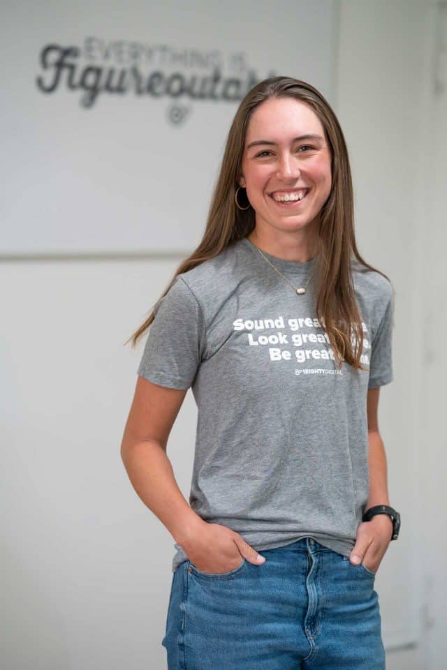 Person in gray tee and denim skirt standing indoors, smiling at website hosting camera.