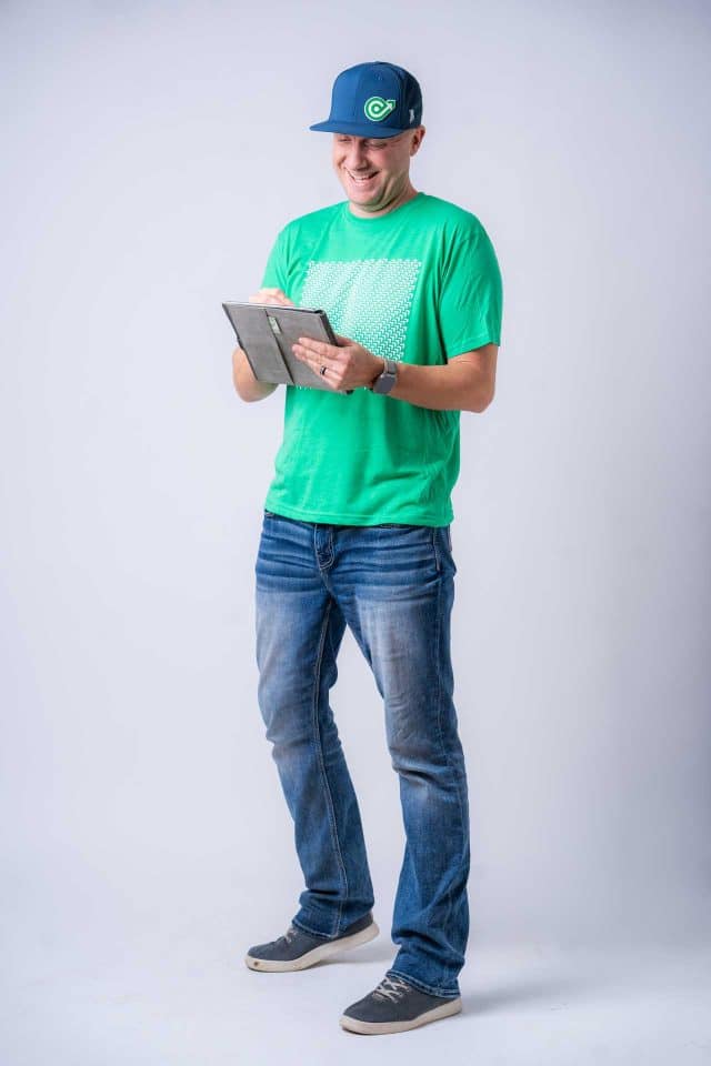 Man in green shirt and jeans writing on a tablet, standing and smiling.