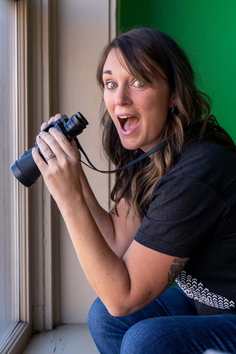 Person smiling, holding binoculars while kneeling by a window.