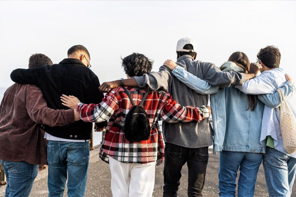 Six people stand side by side with arms around each other, facing away outdoors.