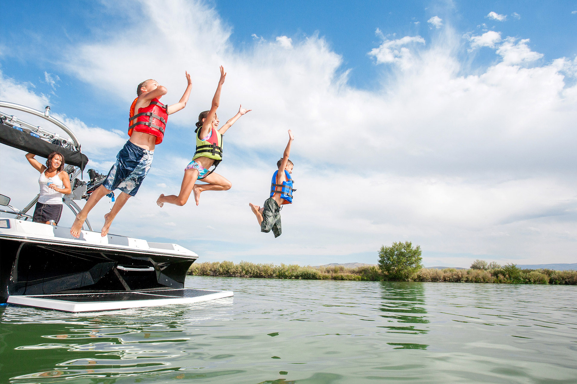 Three people in life jackets jump off a boat into a lake on a sunny day.