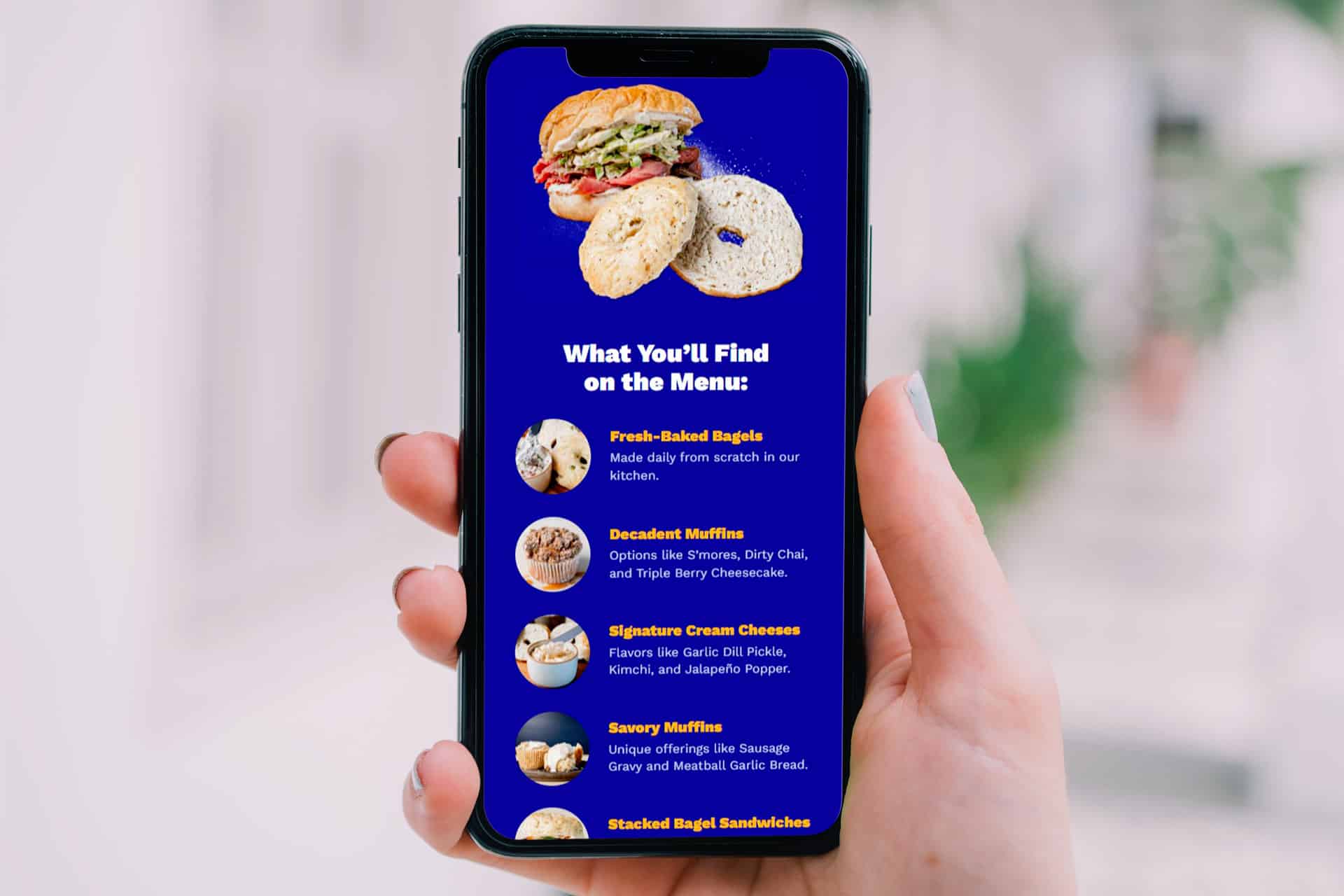 A hand holds a phone displaying a menu with bagels, muffins, and sandwiches.