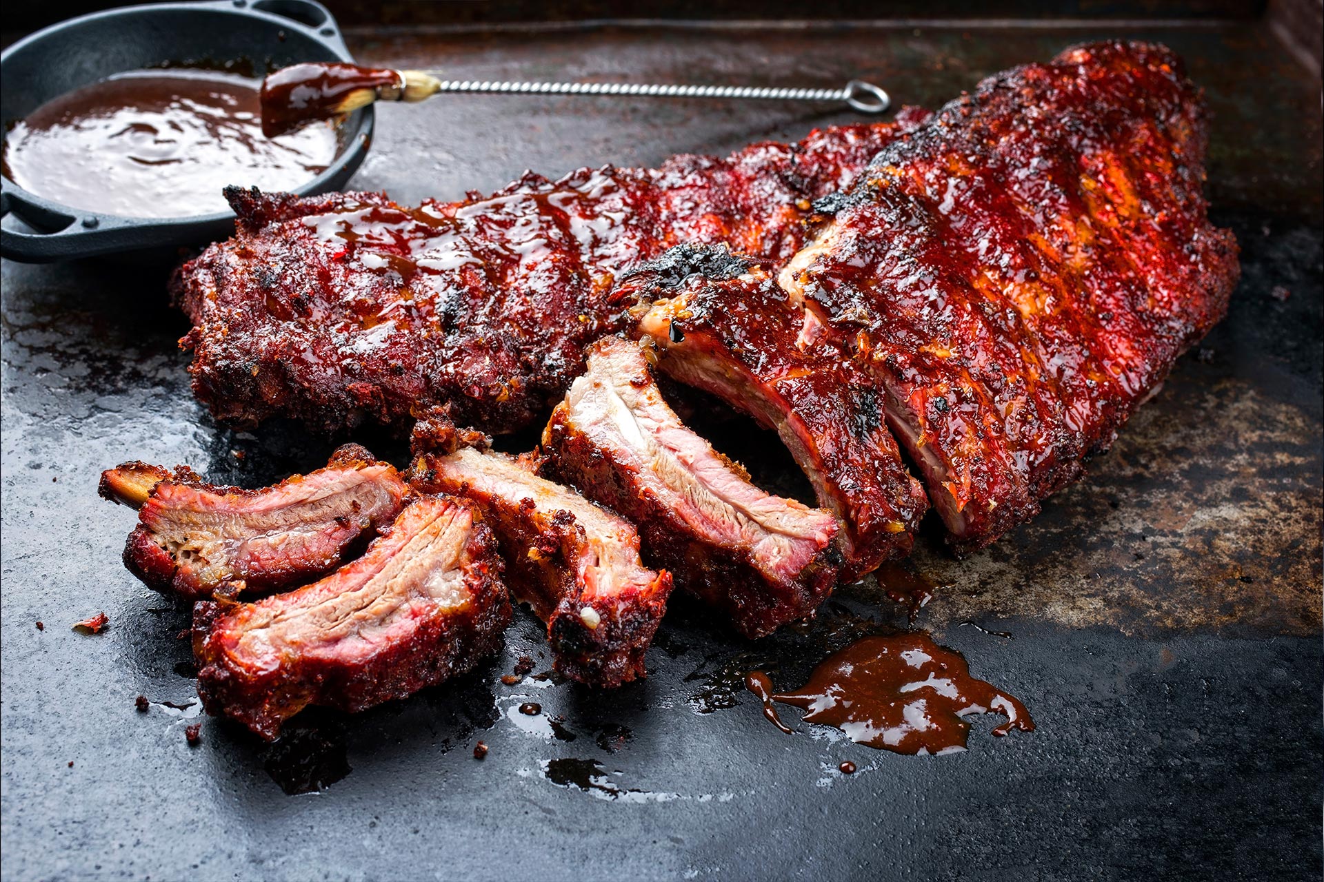 A rack of barbecued ribs with sauce and several ribs sliced on a dark tray.