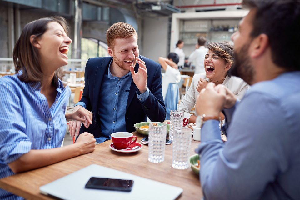 Four people laugh together while sitting at a café table with drinks and food.