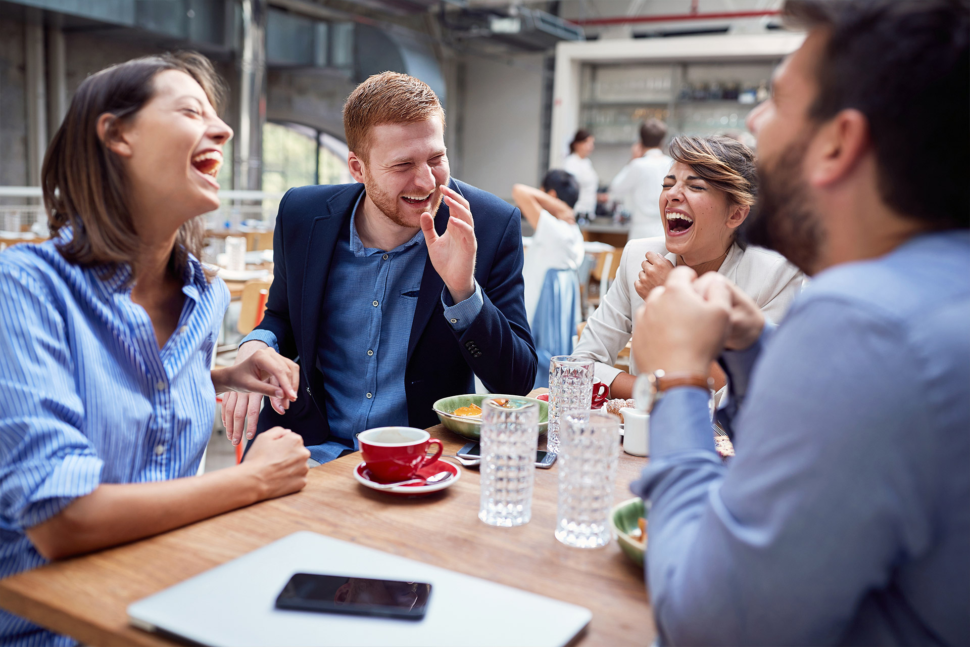 Four people laugh together while sitting at a café table with drinks and food.