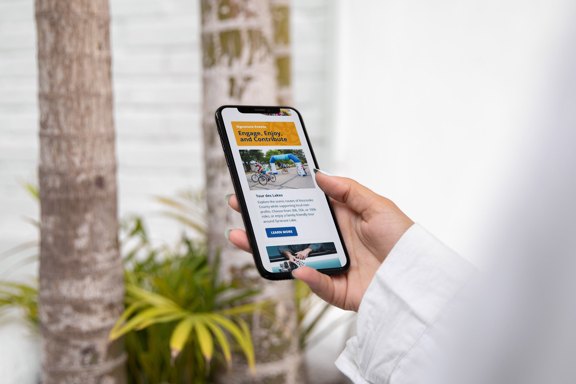 A person holding a smartphone displaying a website outdoors near palm trees.