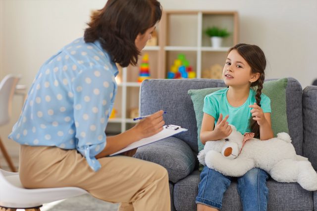 Woman taking notes while talking to a girl holding a stuffed animal on a couch.
