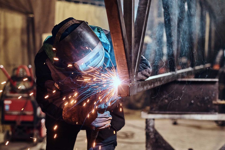 A person welding metal beams with sparks flying in an industrial workshop.