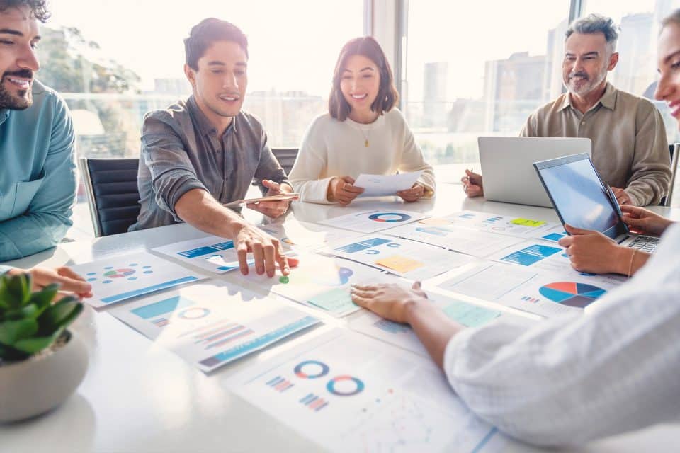 Five people review charts and graphs at a table in a bright office meeting room.
