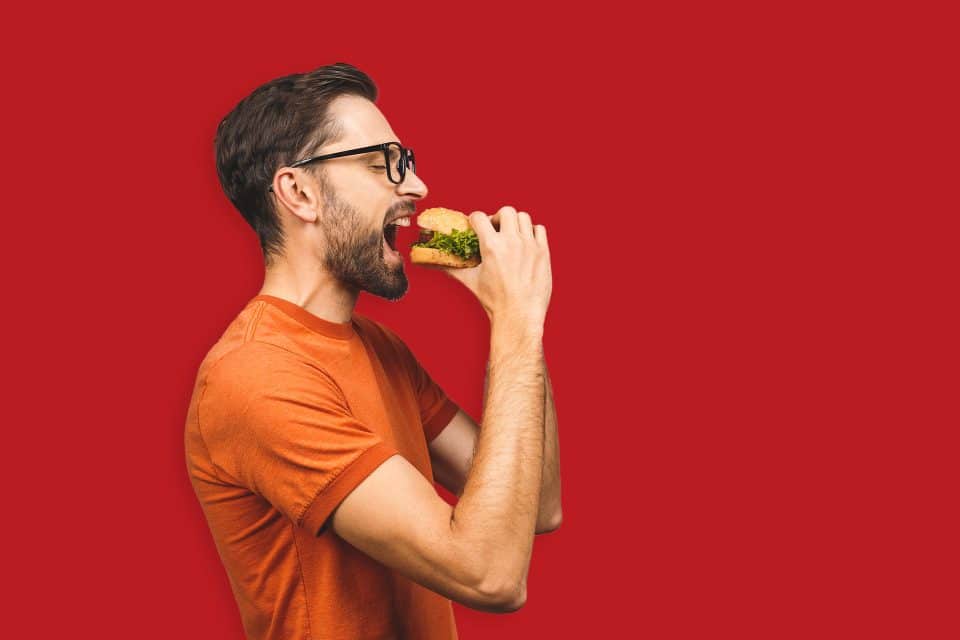 Man in an orange shirt eating a sandwich against a red background.
