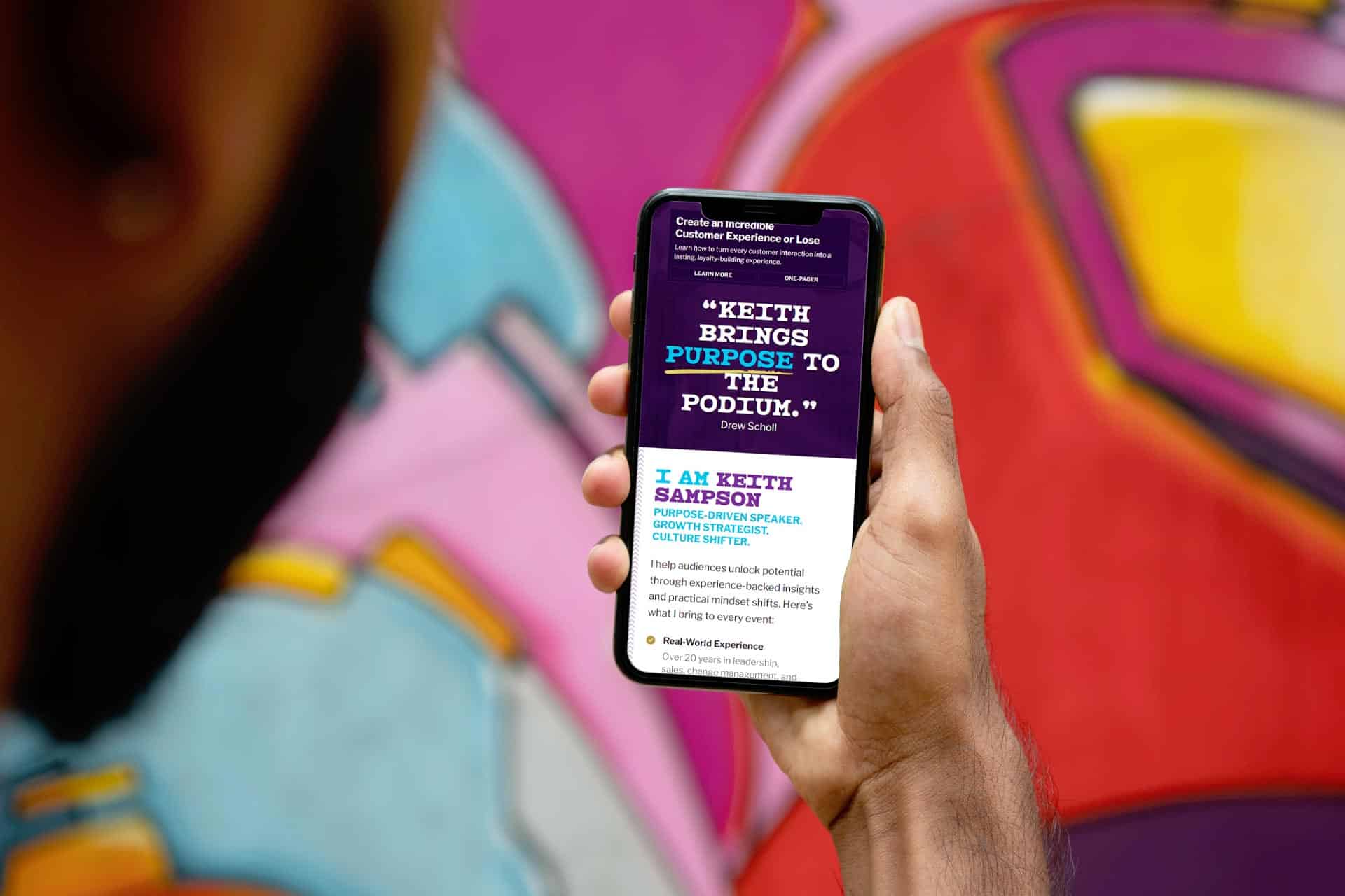 Person holding a smartphone displaying a speaker profile against a colorful mural.