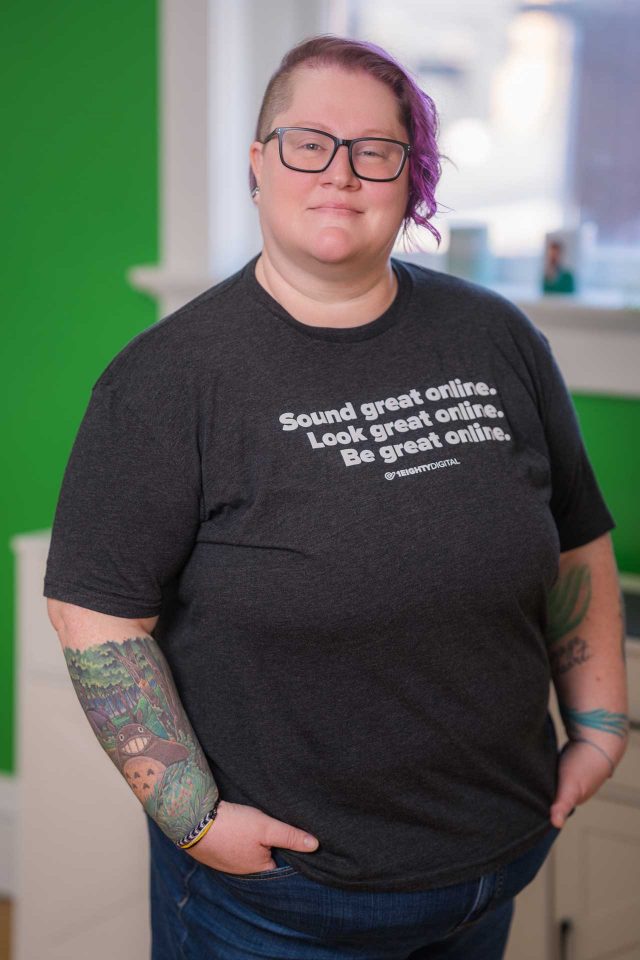Person with glasses and tattoos stands indoors, wearing a printed black t-shirt.