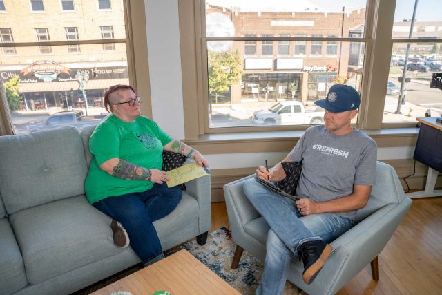 Two people sit on couches in an office, talking and taking notes on clipboards.