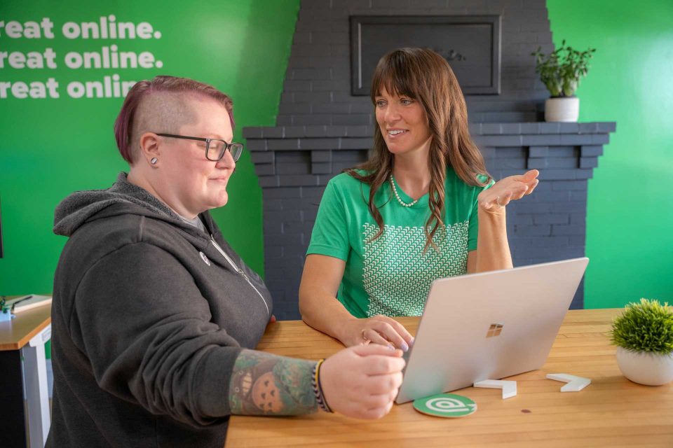 Two people sit at a table with a laptop, conversing in a casual office setting.