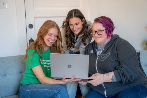 Three people sit on a couch smiling and looking at a laptop screen together.