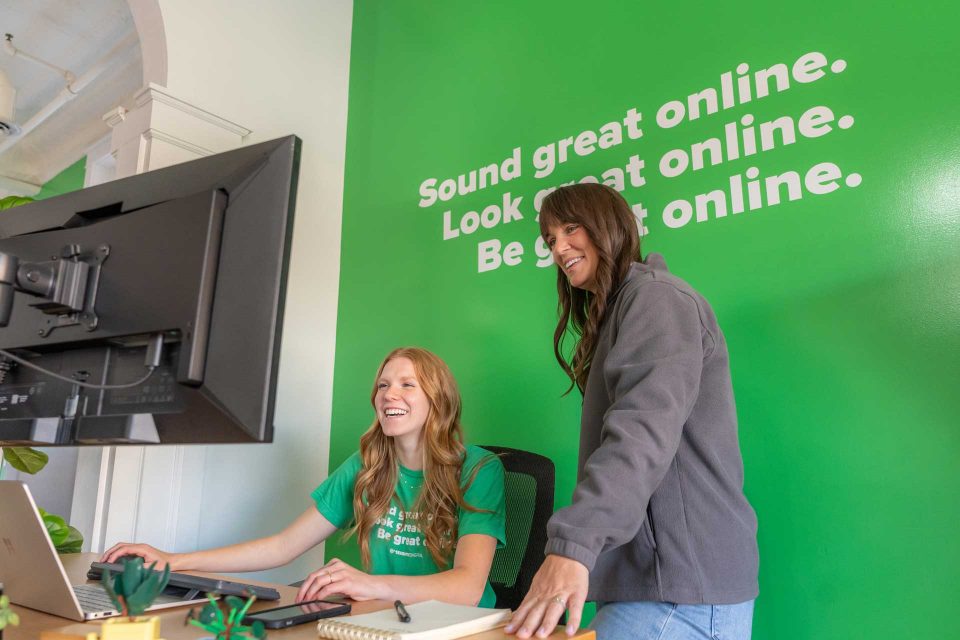 Two women work together at a computer in an office with a bright green wall.