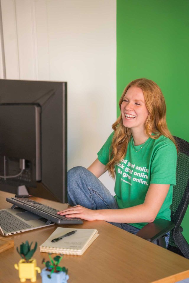 Woman in a green shirt smiling while working on SEO at her computer desk.