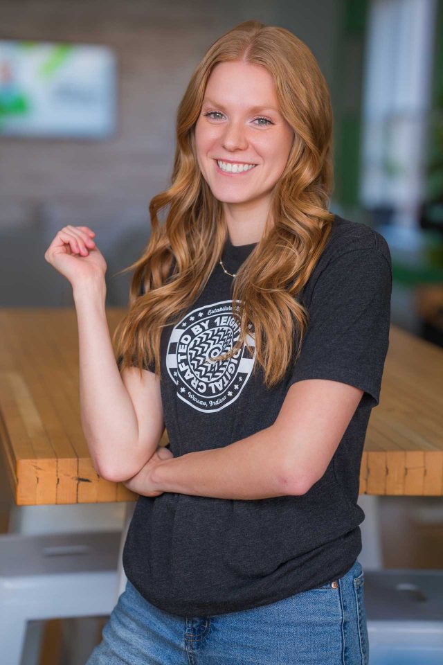 Woman with long, wavy hair smiles, standing indoors by a wooden table.