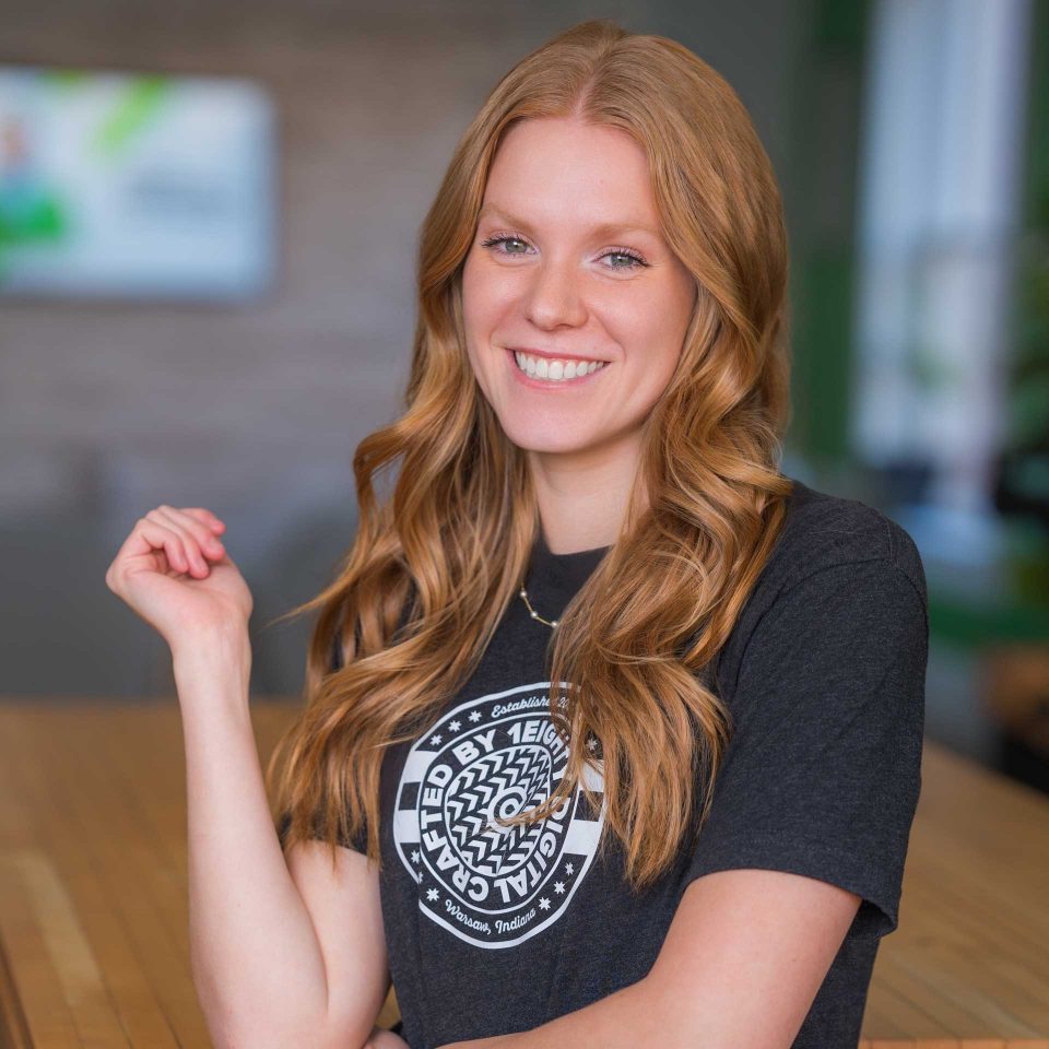 Woman with long red hair smiling, wearing a dark graphic t-shirt indoors.