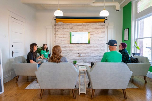 Five people sit in a meeting room, facing a wall-mounted screen displaying data.