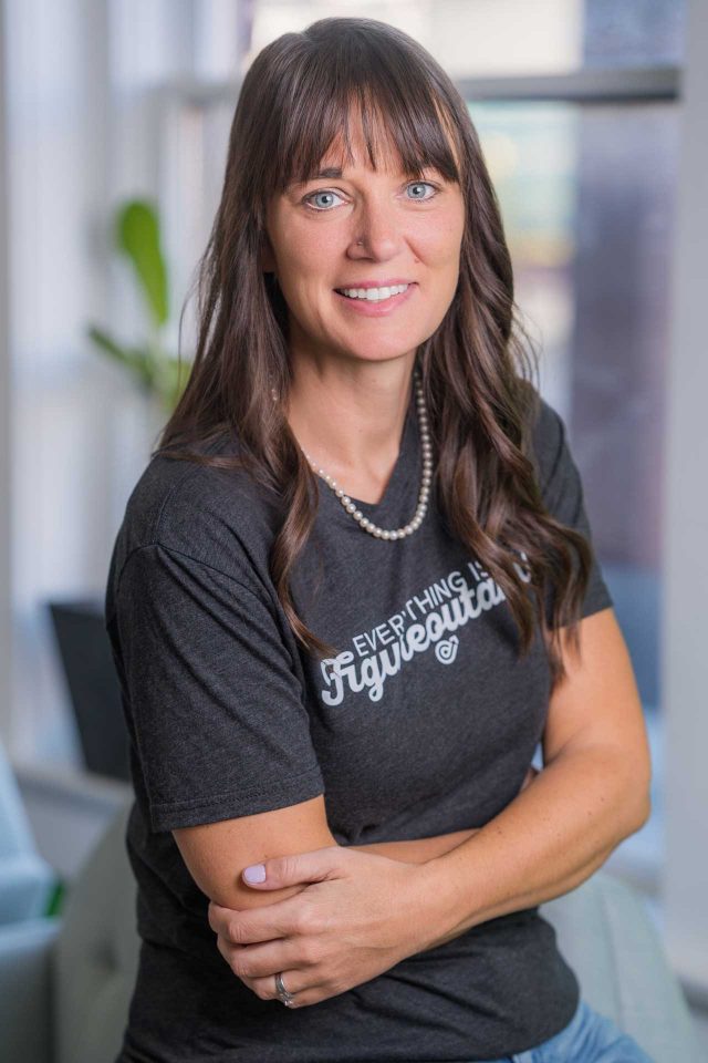 Woman with long brown hair wearing a black t-shirt and a pearl necklace, smiling.