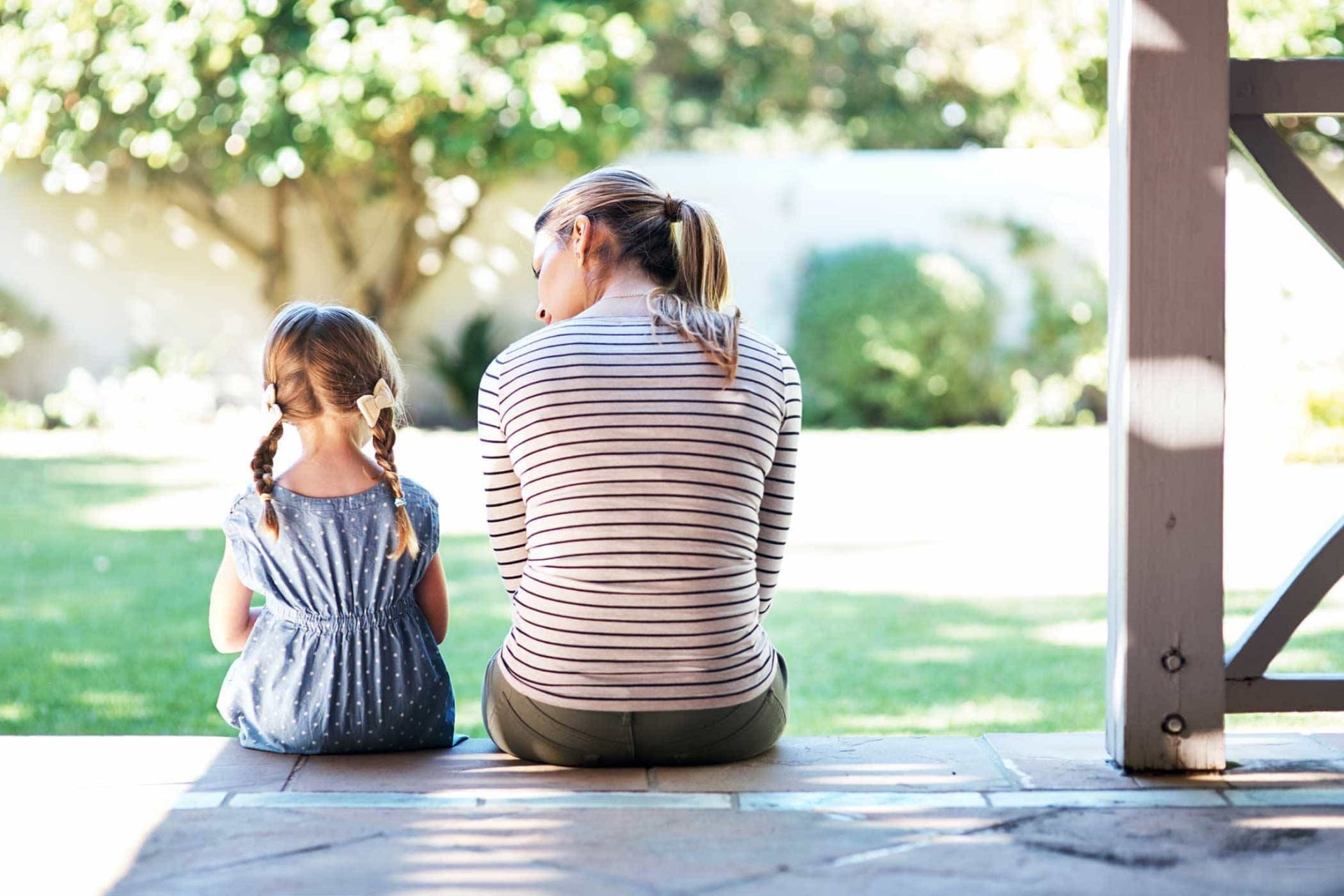 Woman and girl sitting on a porch, facing a sunny backyard.