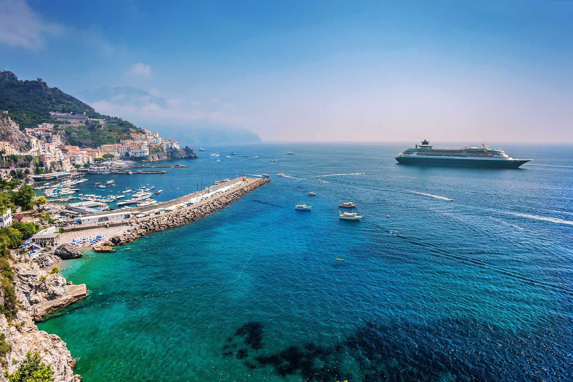 Covington Travel: Cruise ship sails azure seas near coastal town with boats.