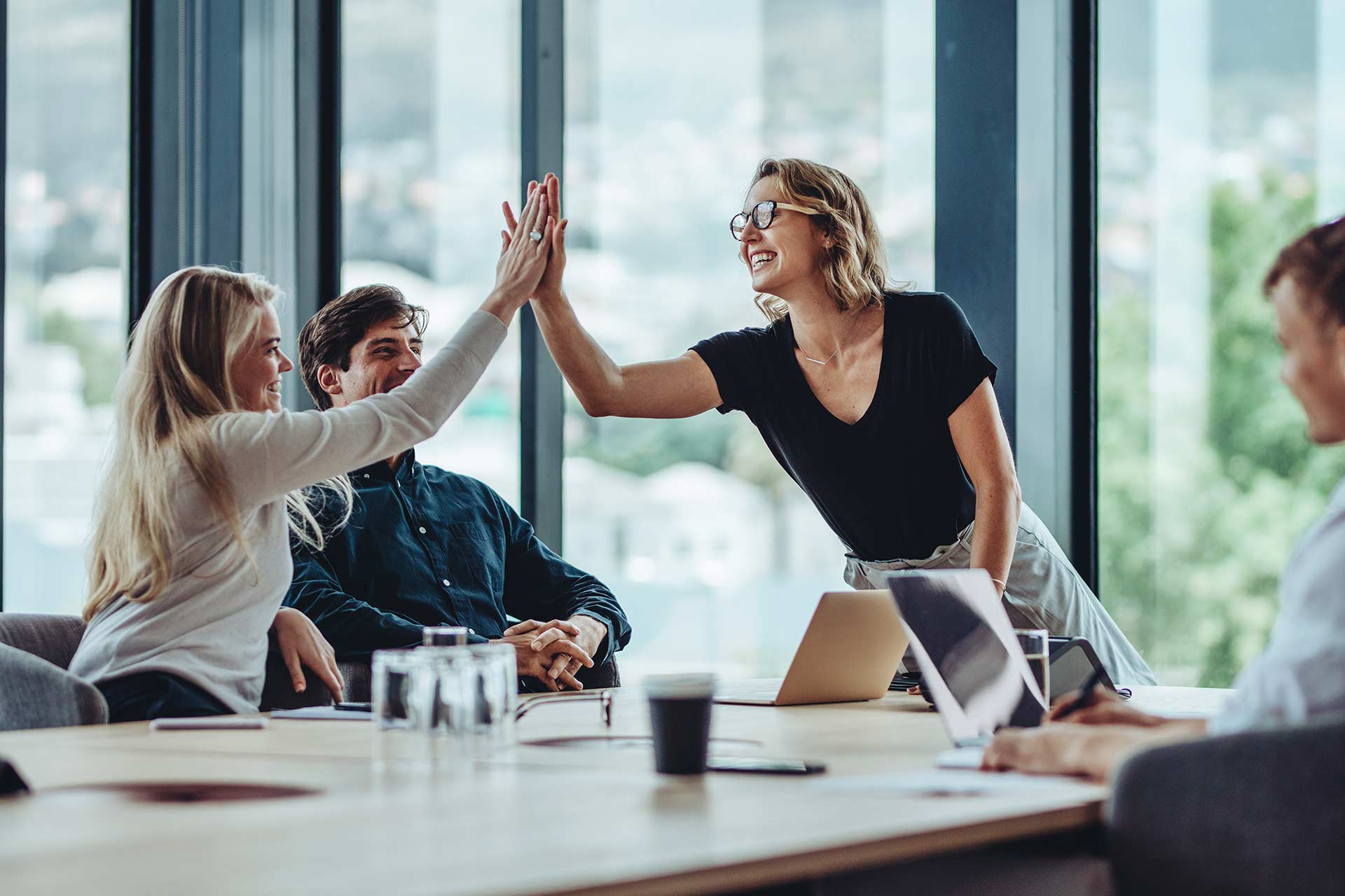 Two women high-five, cheering Dream Again, at a large meeting table with laptops.