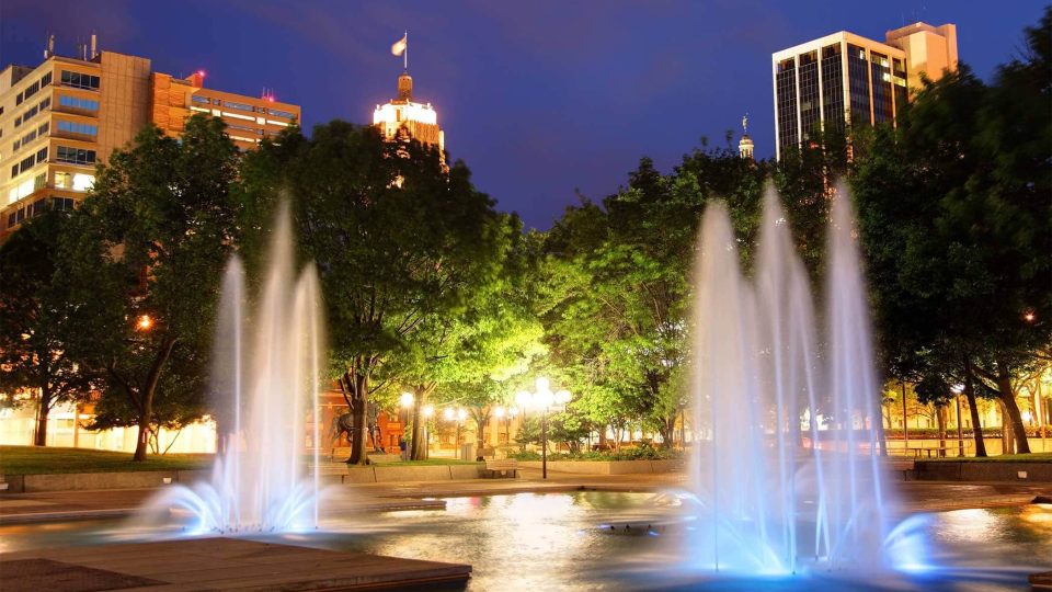 Fort Waynes city park glows with illuminated fountains at dusk.