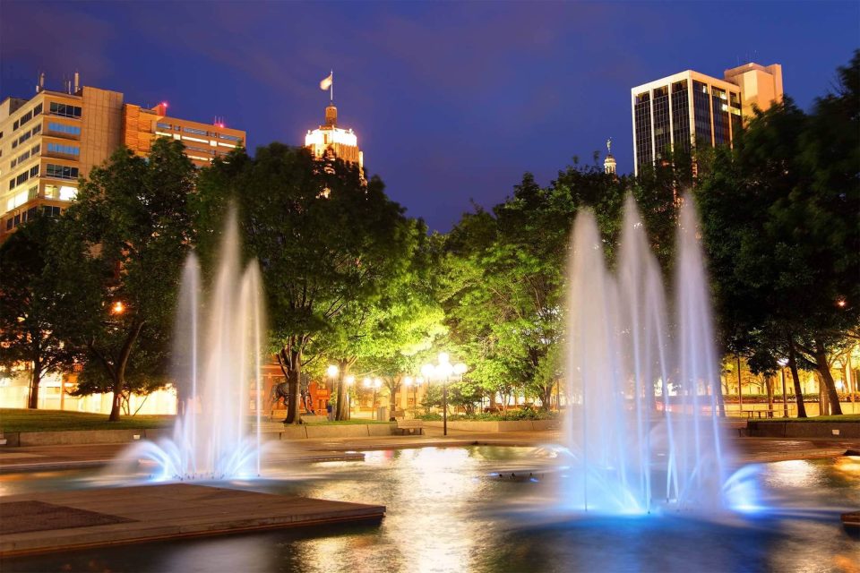 Fort Waynes city park glows with illuminated fountains at dusk.
