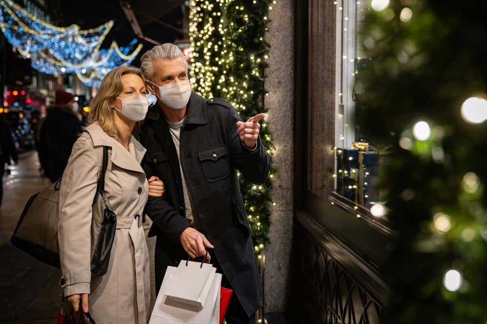 Couple in coats and masks window-shopping amid holiday lights during COVID.