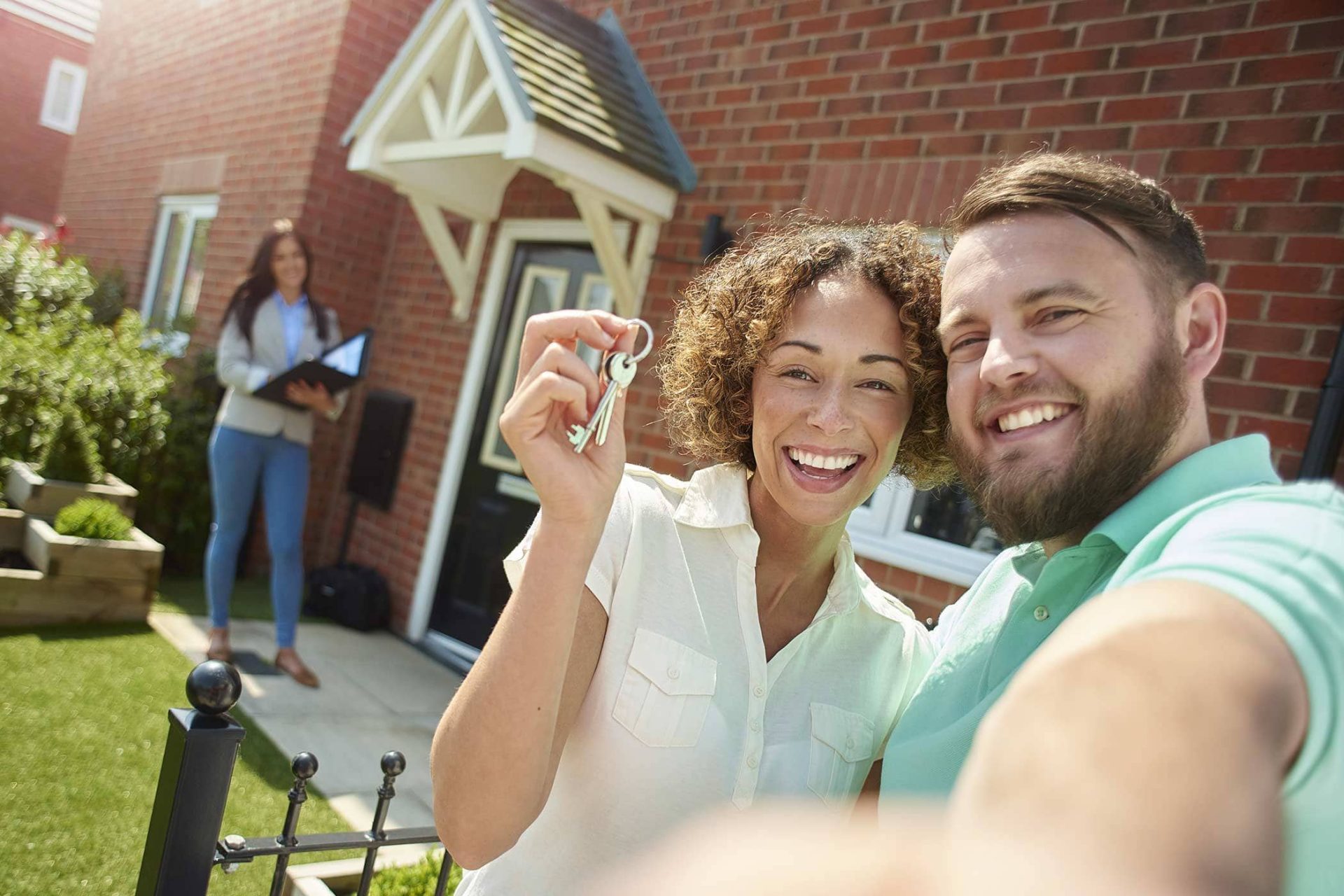 Couple smiling and holding keys in front of new home, a woman stands behind.