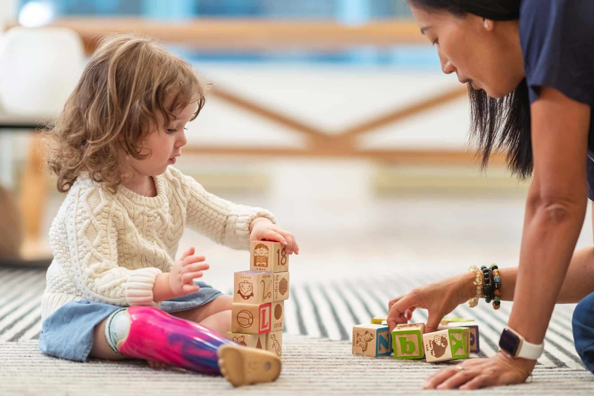 Child with prosthetic leg plays with blocks alongside an adult on a rug.
