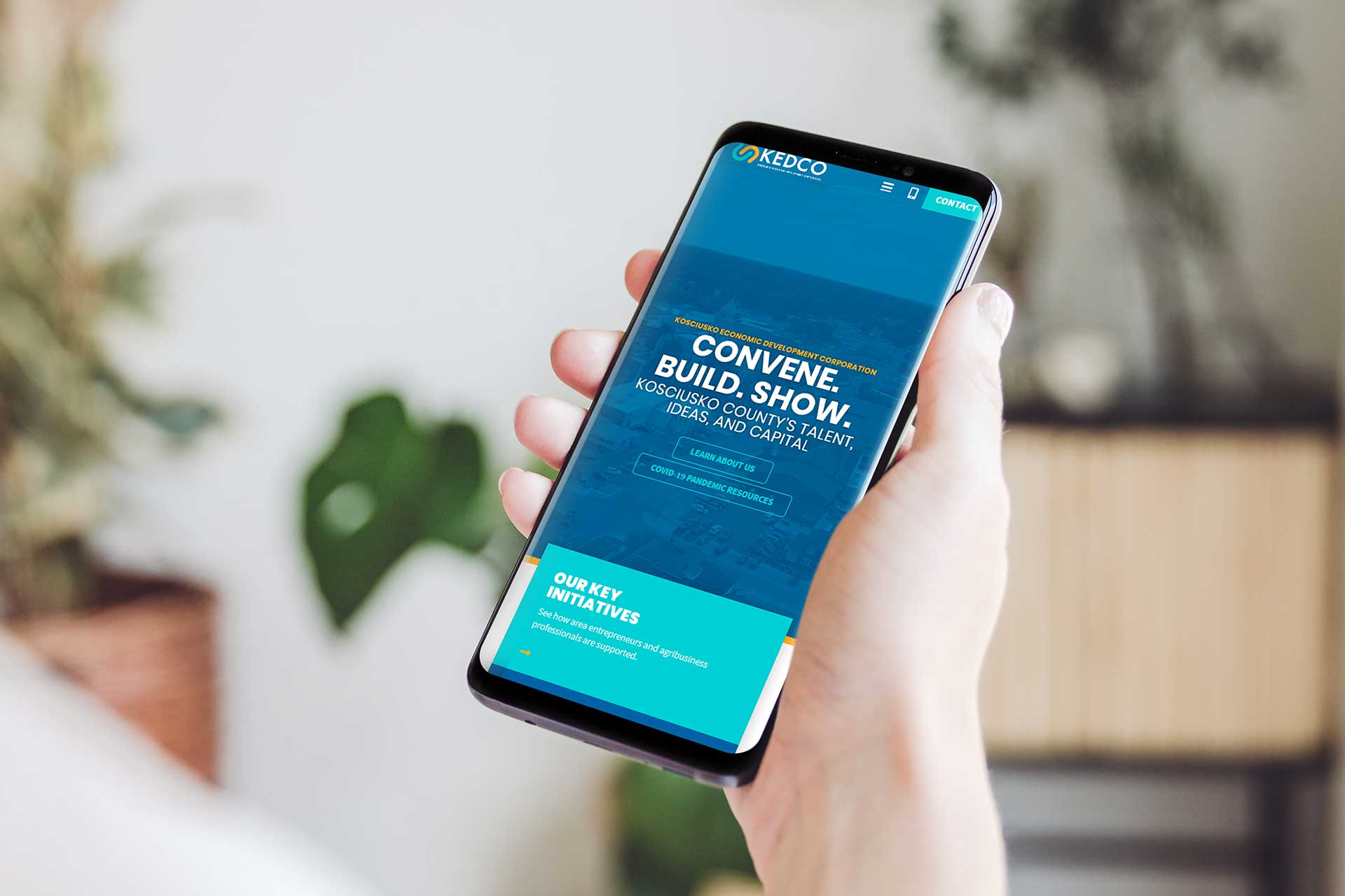Hand holding a smartphone displaying KEDCos website with blue and green text.
