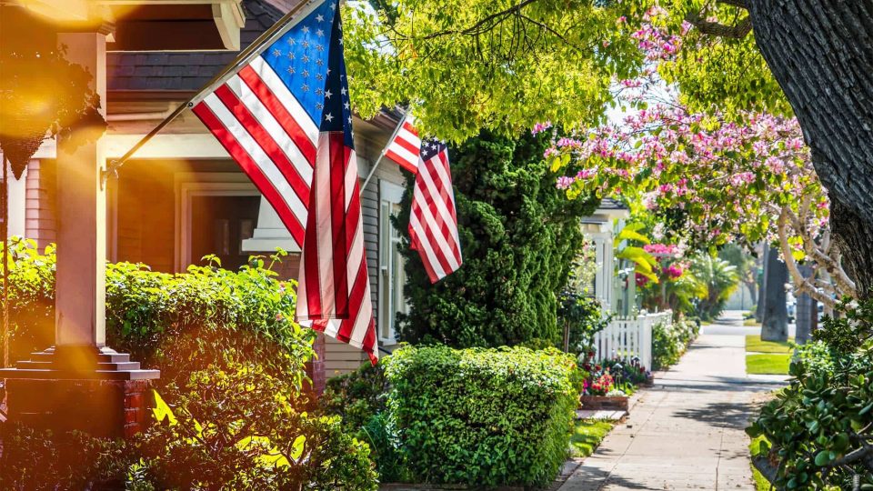 Nappanee homes with flags and greenery line a sunny neighborhood sidewalk.