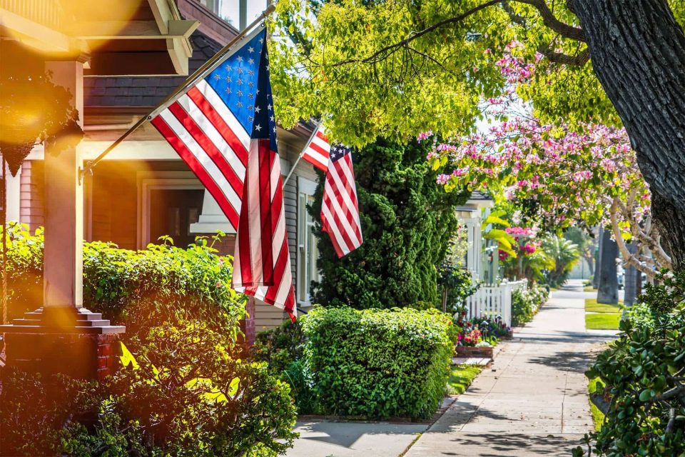 Nappanee homes with flags and greenery line a sunny neighborhood sidewalk.