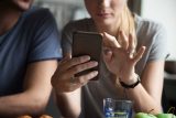 Two people use a smartphone at a table with fruit, water, and email marketing.