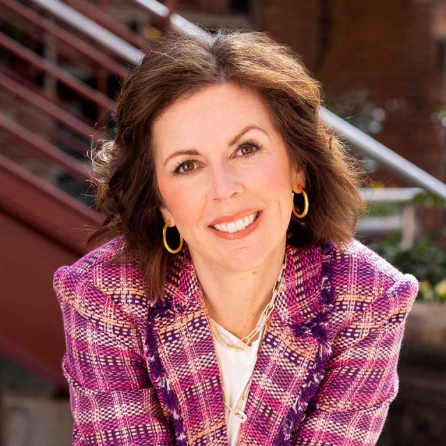 Smiling person in a pink plaid blazer, sitting outdoors.