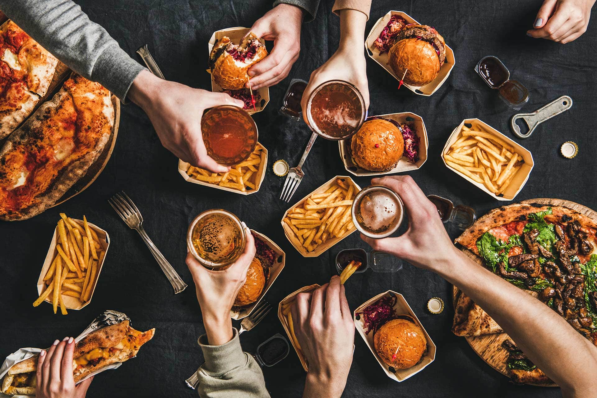 People reaching for burgers, fries, pizza, and drinks on a dark table.