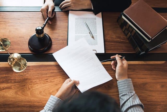 Two people review legal documents at a desk with books, a gavel, and scales.