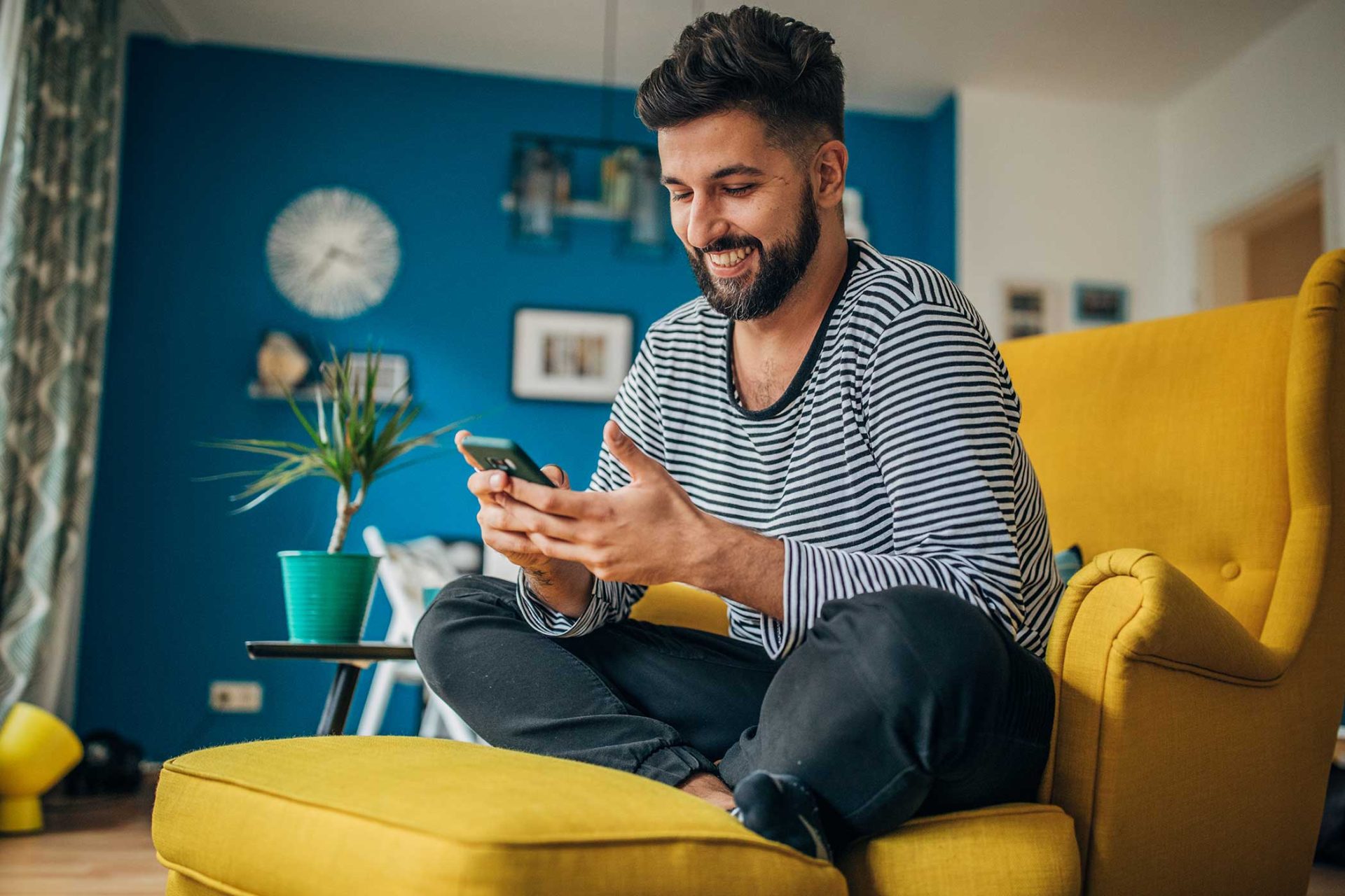 Person sitting on a yellow chair, smiling at a phone in a blue-walled room.