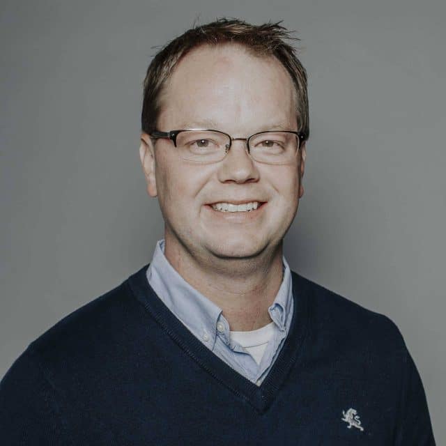 A man wearing glasses and a sweater, smiling against a gray background.