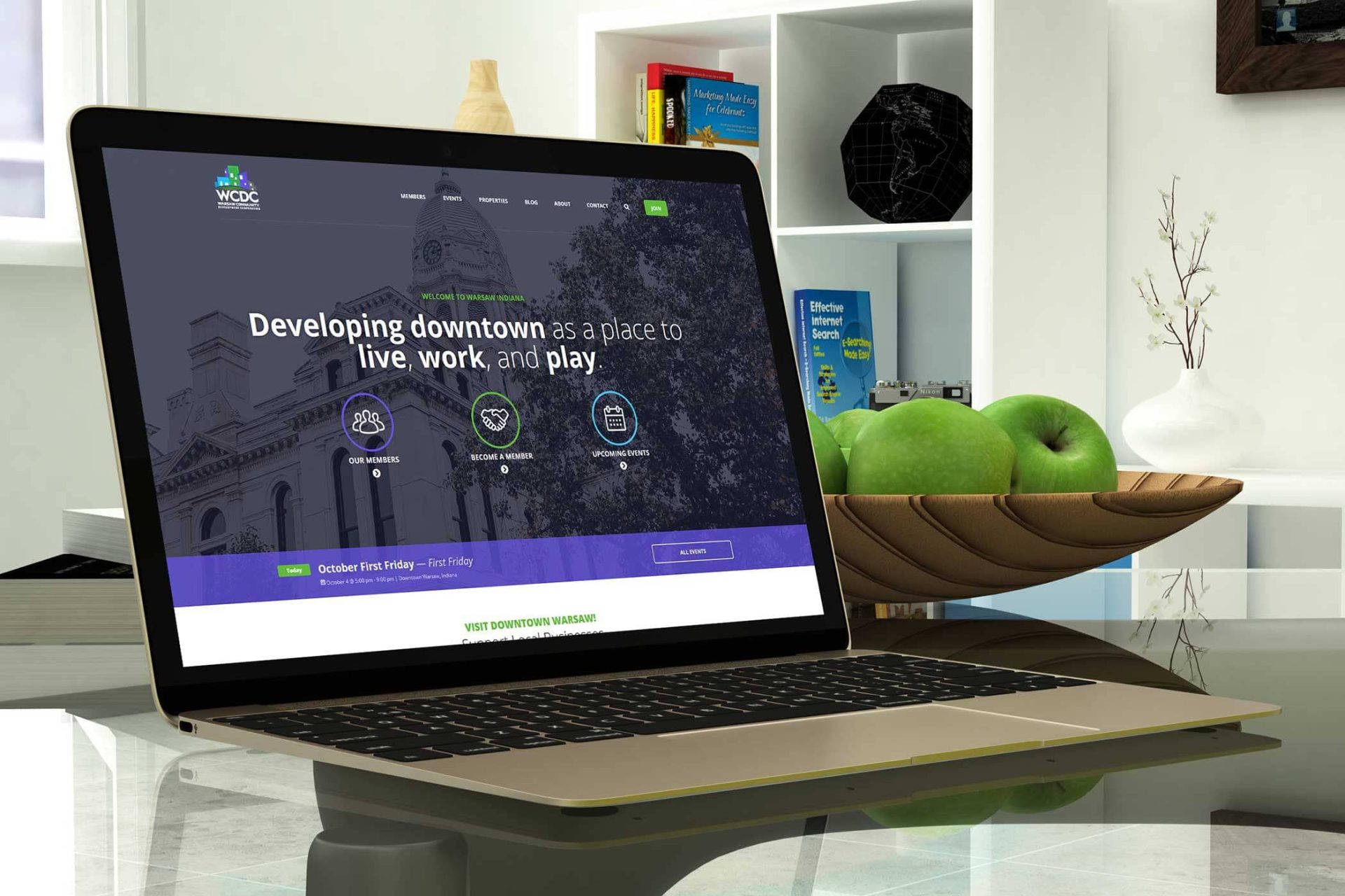 Laptop on desk displaying a website, with green apples and books nearby.