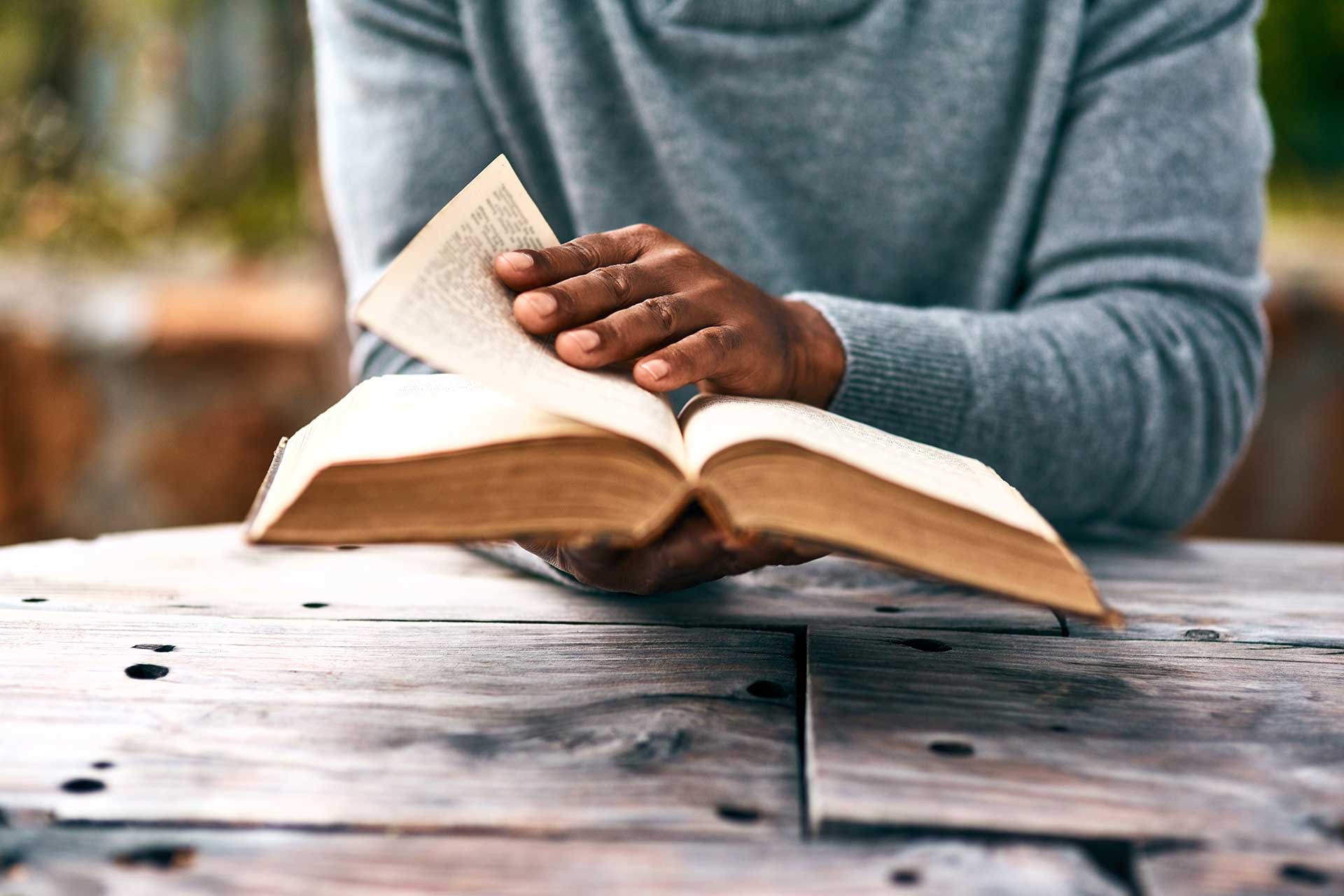 Person reading a book at a wooden table outdoors.