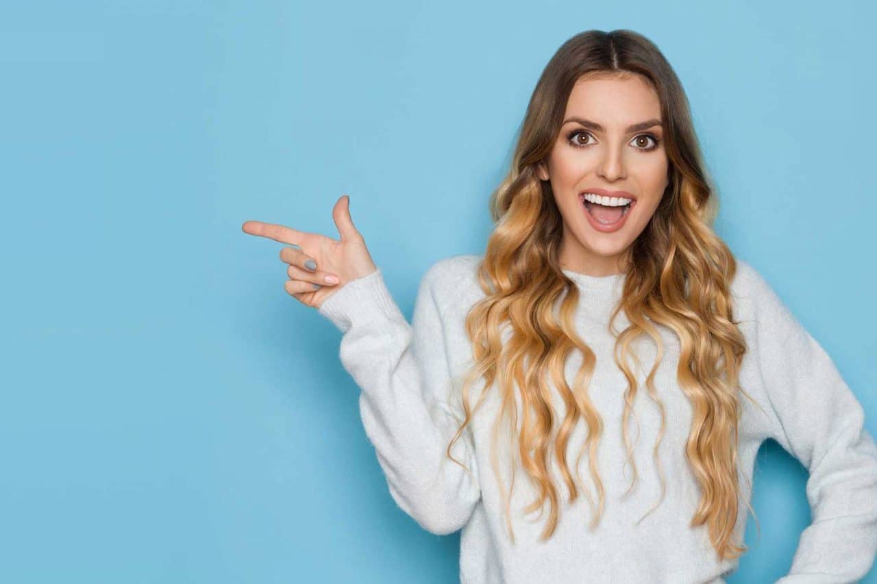 Smiling woman in white sweater pointing to her side with blue background.