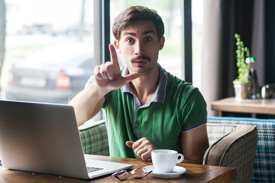 Man gestures loser while seated at cafe table, laptop open to social media followers.